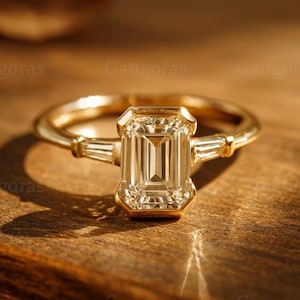 May include: A gold ring featuring a large, rectangular, clear gemstone. Two smaller, rectangular gemstones are set either side of the main stone. The ring is on a wooden surface, with the word "Gangoras" in the background.