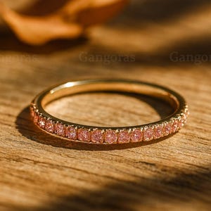 Könnte beinhalten: Ein goldfarbener Ring mit einem Band aus kleinen, rosa Edelsteinen. Der Ring liegt auf einer Holzoberfläche mit verschwommenem Hintergrund. Der Ring ist ein Schmuckstück.