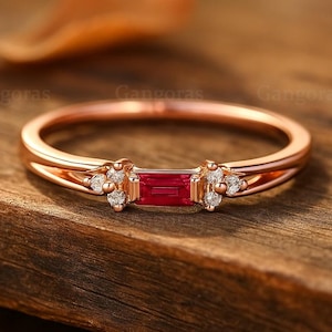 Puede incluir: Anillo de oro rosa con una gema roja rectangular en el centro y pequeños detalles de diamantes. El anillo tiene un diseño de caña dividida y está colocado sobre una superficie de madera. Una joya.