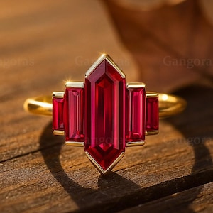 Peut inclure: Une bague en or ornée d'une pierre précieuse rouge foncé rectangulaire centrale, flanquée de pierres rectangulaires plus petites. La bague a un design géométrique et est posée sur une surface en bois.