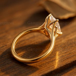 Puede incluir: Un anillo de oro con una gema en forma de pera. La banda y el engaste del anillo son dorados, y la gema es clara y facetada. El anillo está sobre un fondo de madera borroso.