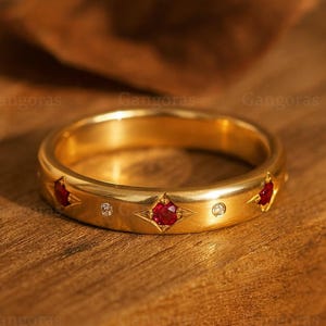 Peut inclure: Une bague en or avec des étoiles rouges en pierres précieuses et de petits diamants clairs. La bague a une finition polie et est posée sur une surface en bois.