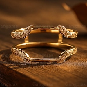 Op de afbeelding: Twee goudkleurige ringversterkers met sierlijke, wervelende patronen. De versterkers zijn ontworpen om rond een ring te zitten en details toe te voegen. De afbeelding is geplaatst tegen een warme, houten achtergrond.