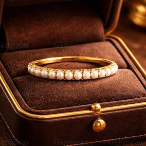 May include: A gold bangle bracelet with a row of white pearls, displayed in a brown jewelry box. The bracelet has a polished gold finish and the pearls are evenly spaced. The jewelry box is brown with a gold trim.