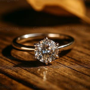 May include: A silver engagement ring featuring a large, round-cut diamond. The diamond is set in a classic six-prong setting. The ring sits on a wooden surface. The ring's design is timeless and elegant.