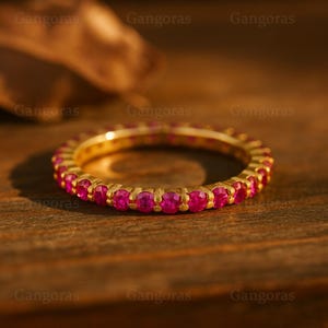 May include: A gold ring featuring a continuous band of bright pink gemstones. The ring is set on a wooden surface, with a blurred background. The gemstones are round and closely set, creating a vibrant line of colour.