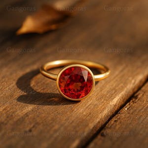 Peut inclure: Une bague en or avec une grosse pierre précieuse rouge et ronde. La pierre est sertie dans un sertissage à lunette, et le bracelet est lisse et poli. La bague est présentée sur une surface en bois.