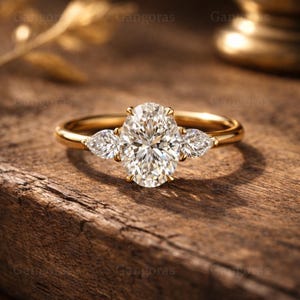 Op de afbeelding: Een gouden ring met een grote ovaal geslepen diamant als middensteen en twee peervormige zijstenen. De ring ligt op een houten oppervlak, waarbij het licht weerkaatst in de facetten van de diamanten. De ring is een sieraad.