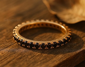 May include: A gold ring featuring a continuous band of small, black gemstones. The ring is set on a wooden surface, with a blurred leaf in the background. The design is simple and elegant, suitable for everyday wear.