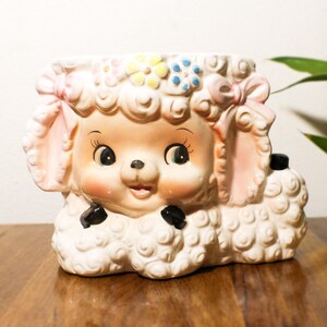 May include: A ceramic lamb planter with a smiling face, large ears, and curly wool. The lamb is painted in soft pink, peach, and white tones, with black accents. It has a floral and bow detail on its head.