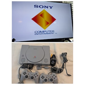 May include: A light gray Sony PlayStation game console with two matching controllers, power and AV cables. The television screen above displays the Sony Computer Entertainment logo with a yellow and red diamond graphic on a white background.