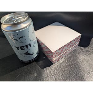 May include: A light blue YETI can cooler featuring black illustrations of a duck and a fish, positioned next to a square white box wrapped in clear plastic with red PENTAX branding printed repeatedly along the edges of the packaging.