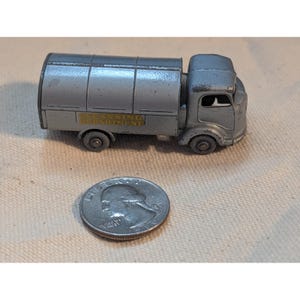 以下が含まれることがあります： 銀色の金属製タンクローリーのミニカー。円筒形のタンクと運転席を備えています。タンクの側面には、黄色い文字で「CLEANSING DEPARTMENT」と書かれた長方形のラベルが貼られています。横にはアメリカの25セント硬貨が置かれています。