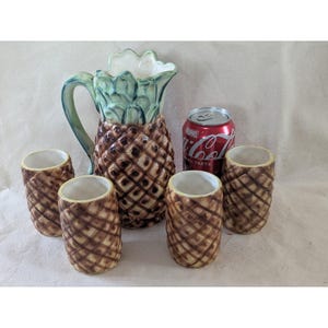 May include: A ceramic pineapple-shaped pitcher with a green handle and top, and a brown body. Four small pineapple-shaped cups and a can of Coca-Cola are next to the pitcher. The can has the word "Marvel" on it.