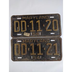 May include: Two rectangular metal Maryland license plates with a black background and raised gold-colored lettering. The top plate reads DD-11-20 and the bottom plate reads DD-11-21. Both plates feature the text 1541 and have four mounting slots.