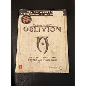 May include: The Elder Scrolls IV Oblivion official game guide with a weathered, off-white cover. The text reads Revised & Expanded, Covers Knights of the Nine, Also Covers PS3, The Elder Scrolls IV Oblivion, Official Game Guide, Covers All Platforms.
