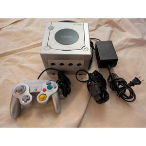 Puede incluir: Consola Nintendo GameCube plateada con un mando plateado a juego. El mando tiene botones de colores y un joystick. También se incluye un adaptador de corriente negro y cables. Se ve el logotipo de Nintendo GameCube.