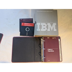 以下が含まれることがあります： ヴィンテージのIBMコンピューターハードウェアとドキュメントの詰め合わせ。グレーのハードカバー本、"Guide to Operations"と書かれたバーガンディ色のバインダー、黒い5.25インチフロッピーディスクがいくつか含まれています。IBMのロゴが複数のアイテムに表示されています。
