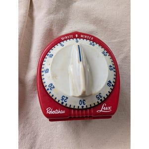 Könnte beinhalten: Ein rot-weißer Küchen-Kurzzeitmesser mit zentralem Ziffernblatt und spitzem Drehknopf. Das Ziffernblatt zeigt blaue Zahlen von 0 bis 55, mit dem Text "MINUTE MINDER" oben und "Robertshaw" sowie "Lux" in Weiß auf der roten Basis.