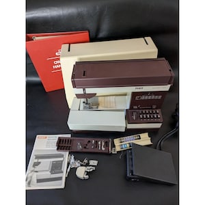 May include: A vintage Pfaff sewing machine in cream and brown, with accessories including a manual, bobbins, and presser feet. The machine has a built-in light and a control panel with stitch options. Accessories are neatly arranged. A red binder is also present.