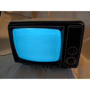 May include: A vintage General Electric television with a white outer casing and a black front panel. The screen displays a bright blue light with horizontal lines. Two circular control knobs are located on the right side, and a thin metal antenna extends from the top.