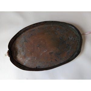 May include: An oval copper tray with a weathered, dark patina and a small metal loop handle on one side. A white measuring tape is visible along the edge, indicating a length of approximately 25 cm for this metallic serving piece.
