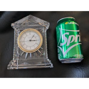May include: A clear crystal clock with a gold-colored bezel and Roman numeral hour markers. The clock is in a triangular-shaped case. A green Sprite soda can is next to the clock.