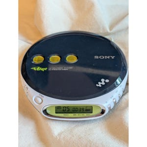 May include: A round, black and white Sony CD Walkman D-EJ360 portable music player with G-Protection. The device features three yellow circular buttons on the top, a digital LCD screen displaying track 05 and time 00:39, and a white side panel.