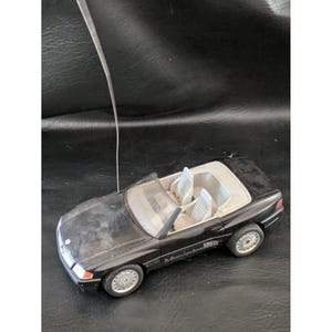 May include: A black remote-controlled Mercedes 500 SL convertible toy car featuring a light gray interior, a clear windshield, silver-colored wheels, and a long, thin metal antenna extending upward from the hood of the vehicle.