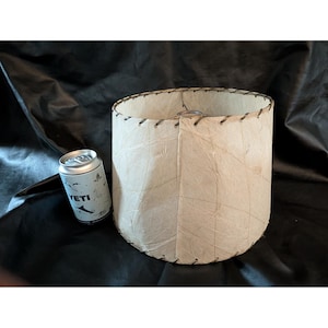 May include: A cylindrical lamp shade with a textured, off-white, fibrous surface and dark stitching along the top and bottom edges. A silver-colored YETI beverage can is positioned next to the lamp shade to provide a visual scale reference.