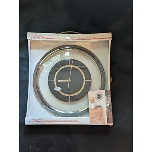 May include: A Sunbeam round wall clock in original packaging. The clock features a black circular face with gold-tone accents, gold hands, and a clear glass lens. The packaging includes the Sunbeam logo and text reading Precision Quartz Accurate Timekeeping.