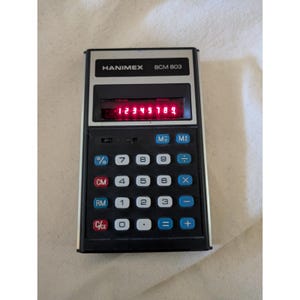 May include: A vintage Hanimex BCM 803 electronic calculator with a black and silver rectangular body. The device features a dark display screen showing the red illuminated numbers 1 2 3 4 5 6 7 8 9 and a grid of square buttons with white and blue labels.