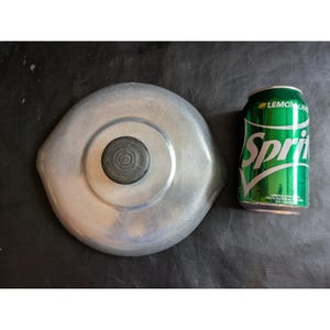 Op de afbeelding: Een ronde deksel van geborsteld aluminium met een centrale, ronde zwarte knop met een textuur van concentrische ringen. De deksel ligt naast een groen Sprite-blikje van 355 ml voor een visuele vergelijking van de grootte.
