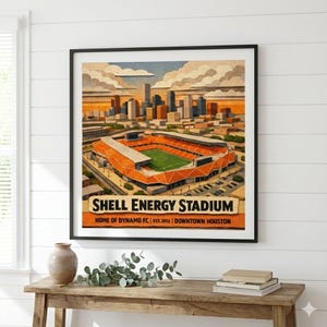 May include: Framed art print of Shell Energy Stadium, home of Dynamo FC in downtown Houston. The artwork depicts an orange and green stadium with a cityscape backdrop under a cloudy sky. The print is displayed on a wooden table.