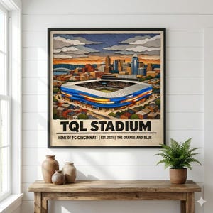 Puede incluir: Una impresión artística enmarcada del estadio TQL, sede del FC Cincinnati, con una vibrante ilustración del estadio y el paisaje urbano circundante. La obra de arte presenta el texto "TQL STADIUM" y "HOME OF FC CINCINNATI | EST. 2021 | THE ORANGE AND BLUE".