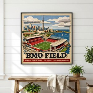 May include: A framed art print of BMO Field, home of Toronto FC, featuring a stadium with a green field and red seating. The artwork includes a cityscape with the CN Tower, water, and clouds.