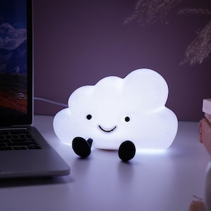 Peut inclure: Une veilleuse en forme de nuage blanc avec un visage souriant et des pieds noirs. La lampe brille et le design est simple et mignon. Elle est posée sur une surface blanche.