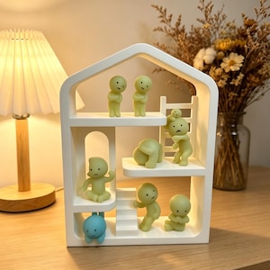 May include: A white, multi-tiered shelf in the shape of a house, populated by small, light green figurines in different poses. A blue figurine hangs from the bottom. The scene is lit by a warm light, creating a cosy atmosphere.
