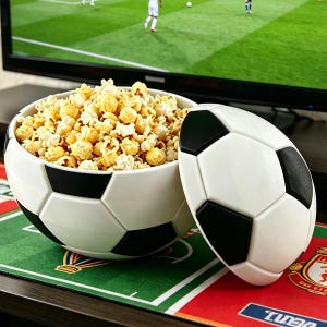 Könnte beinhalten: Eine weiß-schwarze Schüssel in Fußballform, gefüllt mit Popcorn, mit passendem Deckel. Die Schüssel steht auf einer grün-roten Oberfläche, mit einem Fernseher, der ein Fußballspiel im Hintergrund zeigt.