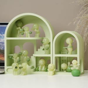 Puede incluir: Una estantería decorativa verde claro de varios niveles con aberturas arqueadas. En el interior, pequeñas figuras brillantes están dispuestas en varias poses, algunas con accesorios como un tambor, pesas y un hula hoop. Las figuras son de color amarillo pálido.