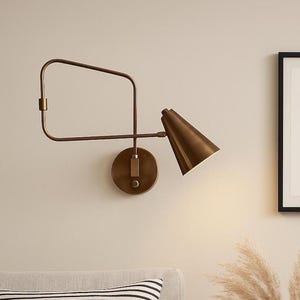 May include: A bronze wall sconce featuring a conical lampshade and a rectangular arm. The sconce is fixed to a round base and set against a neutral wall. The light is on, casting a glow on the wall.