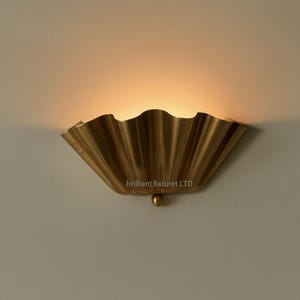 May include: A brass-colored wall sconce with a scalloped, fan-like design. The fixture has a small, round detail at the bottom and the text "brilliant fixturet LTD" is visible. The light shines from behind the scalloped edge.
