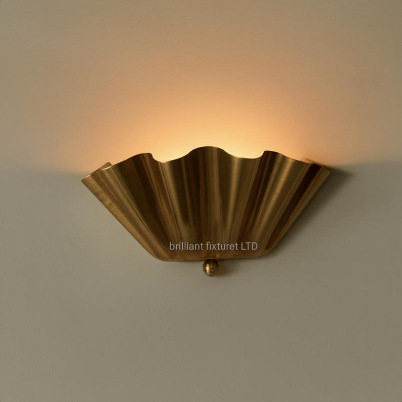 May include: A brass-colored wall sconce with a scalloped, fan-like design. The fixture has a small, round detail at the bottom and the text "brilliant fixturet LTD" is visible. The light shines from behind the scalloped edge.