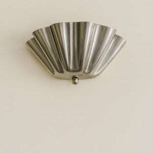 May include: A silver-colored, scalloped wall sconce with a small, round base. The sconce has a fan-like design with multiple curved sections. The fixture is mounted on a light beige wall, creating a contrast in color and texture.