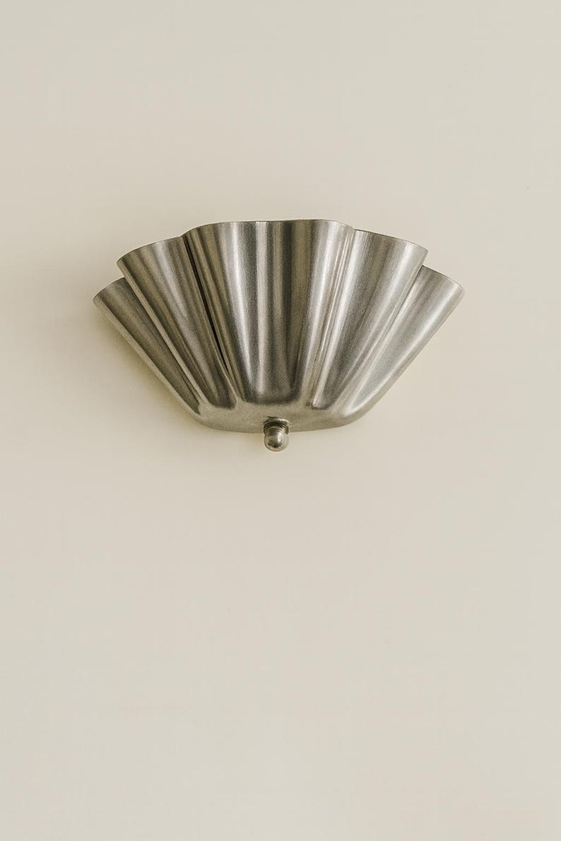 May include: A silver-colored, scalloped wall sconce with a small, round base. The sconce has a fan-like design with multiple curved sections. The fixture is mounted on a light beige wall, creating a contrast in color and texture.