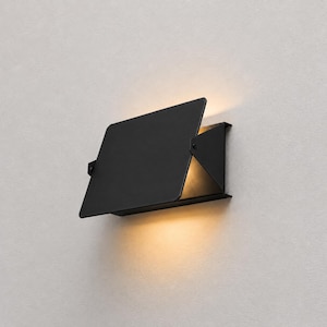 Pode incluir: Um candeeiro de parede preto mate com uma placa frontal retangular inclinada que oculta parcialmente a fonte de luz. A luminária emite um brilho quente e suave pelas bordas superior e inferior, criando um padrão de luz focado na parede.