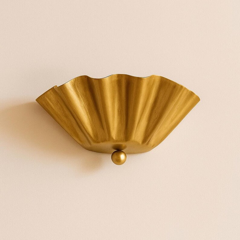May include: A gold-colored, wall-mounted decorative bowl with a scalloped edge. The bowl has a textured surface and a small, round, gold-colored ball at the bottom. The bowl is mounted on a light-colored wall.