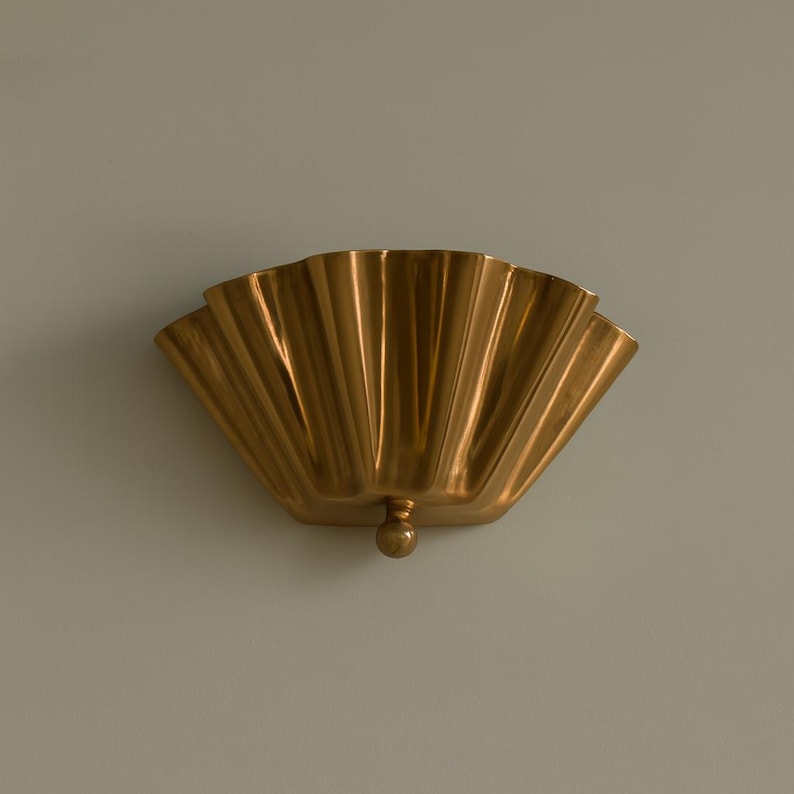 May include: A gold-colored, fan-shaped wall sconce. The sconce has a scalloped design with vertical ridges and a small, round decorative element at the bottom. The fixture is mounted on a light gray wall.