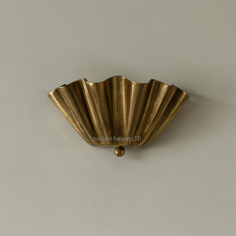 May include: A brass-colored wall sconce with a scalloped, fan-like design. The sconce has a textured surface with vertical lines and a small, round detail at the bottom. The sconce is mounted on a light-colored wall.