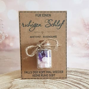 Könnte beinhalten: Eine kleine Glasflasche gefüllt mit Amethyst- und Rosenquarz-Kristallen, mit Bindfaden gebunden, liegt auf einer braunen Karte. Die Karte hat deutschen Text: "Für einen ruhigen Schlaf" und "Falls der Kopf mal wieder keine Ruhe gibt."